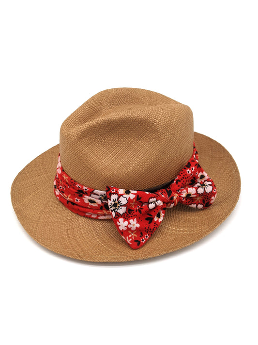 Brown panama hat rests on a white backdrop. The fedora style hat has a red and white flowery band around the middle with a large bow.